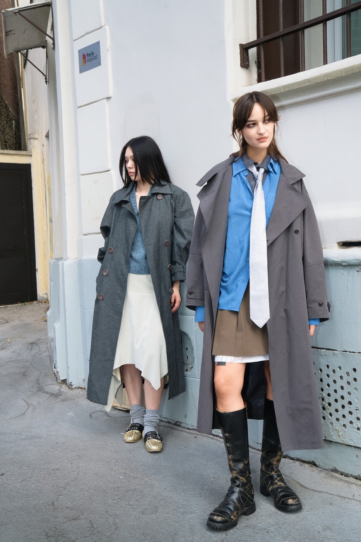 Model on the left wearing the MIST AW26 grey belted double-breasted trench coat outdoors.